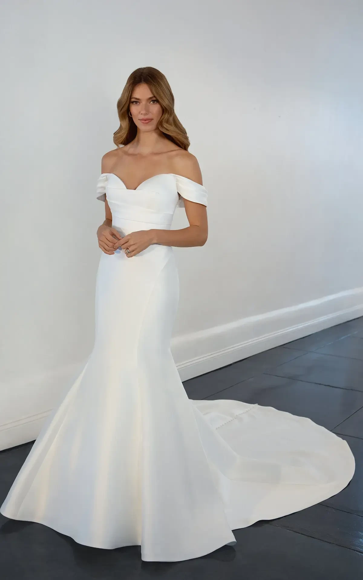 A woman in an elegant, off-shoulder white wedding gown stands against a plain light gray wall. The dress features a fitted bodice and a flowing train.