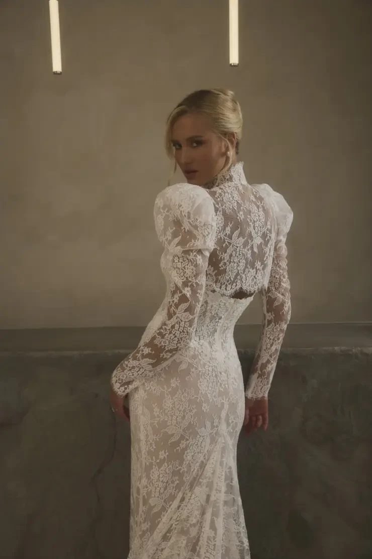 A woman in a strapless, lace wedding gown stands elegantly in a neutral-toned room. The dress has a fitted silhouette and a long train, enhancing the serene and sophisticated ambiance.