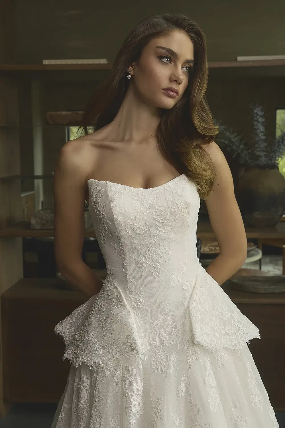 A model wearing a strapless white wedding dress with intricate lace detailing and a peplum design, posing in an elegantly decorated room.