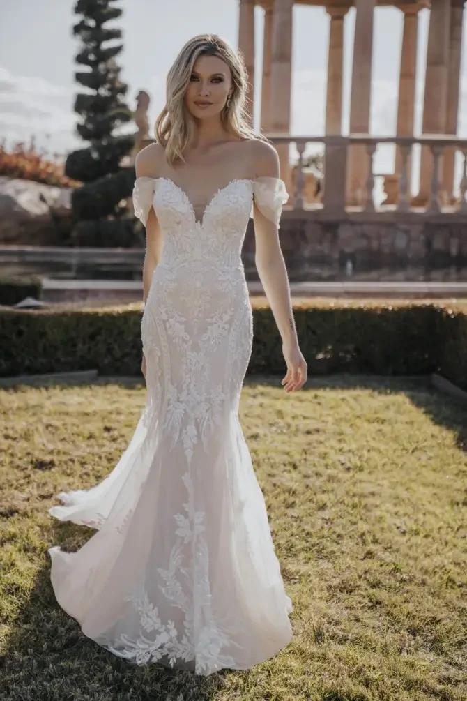 A woman in an off-the-shoulder wedding dress stands in a garden with classical architecture in the background.