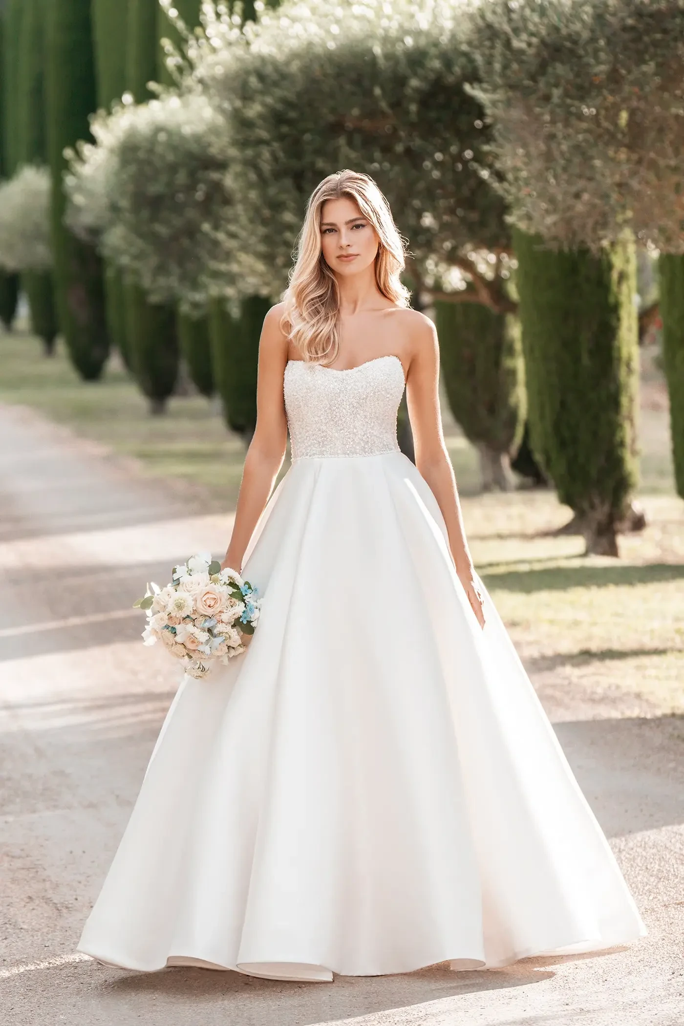 Bride in a strapless white gown, standing on a sunny path lined with cypress trees. Holds a bouquet of white and pastel flowers, exuding elegance and serenity.