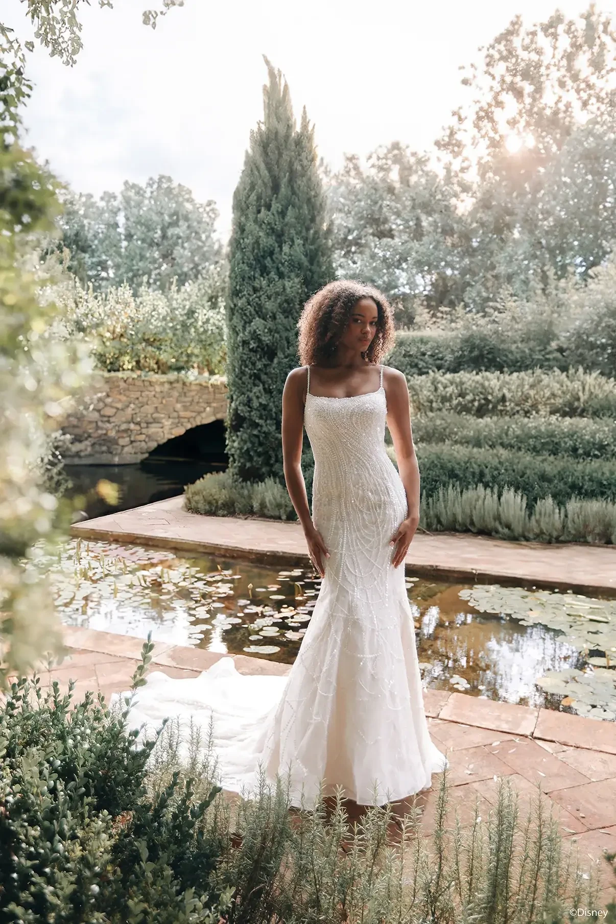 A woman in a fitted white wedding dress stands gracefully in a garden setting, with greenery and water features in the background.