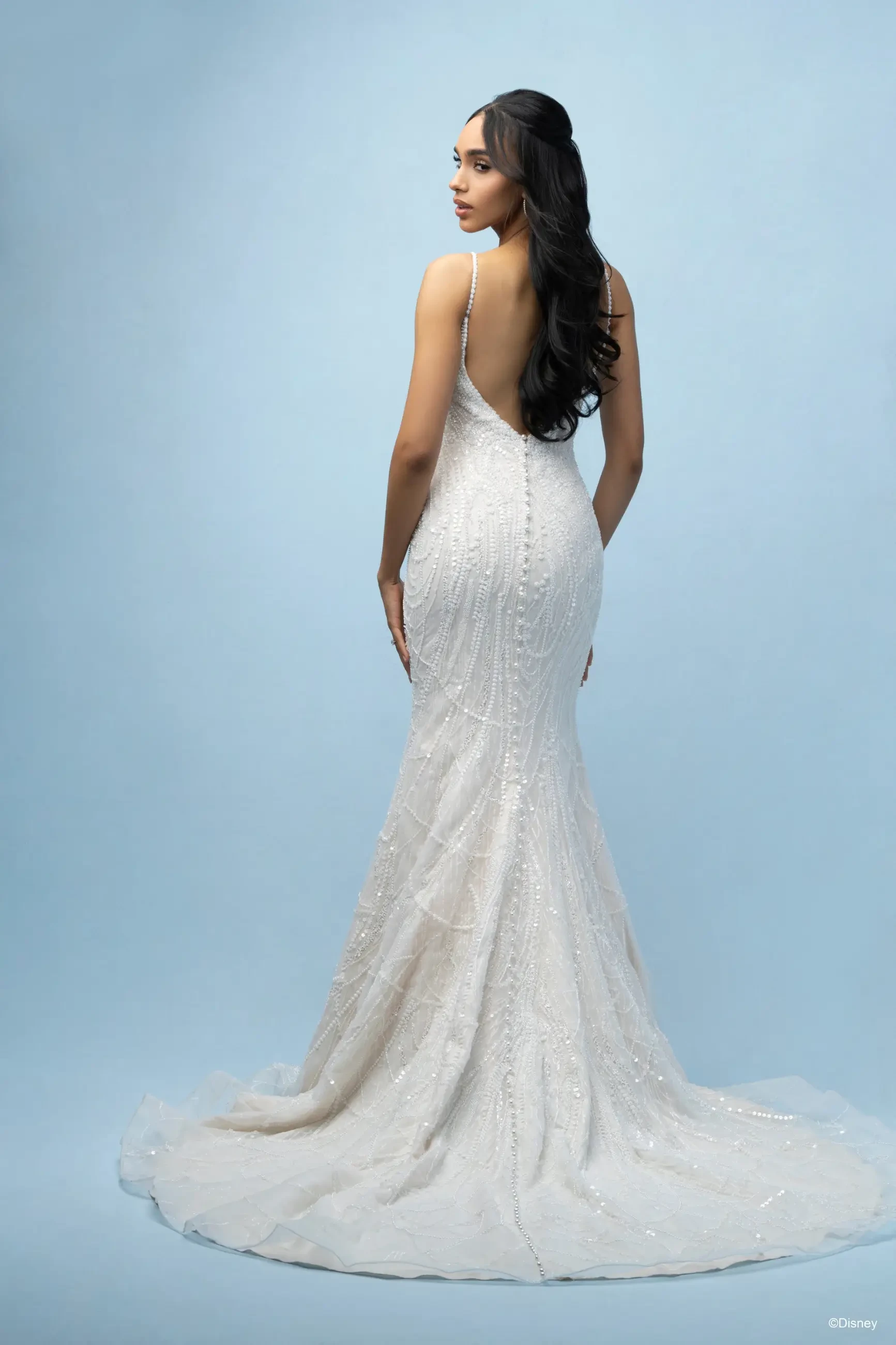 A woman wearing a form-fitting white gown, seen from the back against a light blue background. The gown has intricate details and a train.