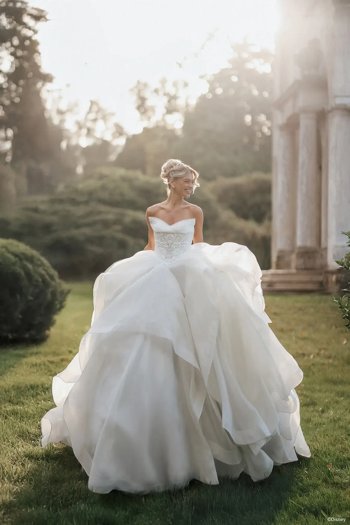 A bride in a stunning white wedding dress with a voluminous skirt stands in a garden, smiling softly as sunlight filters through the trees.
