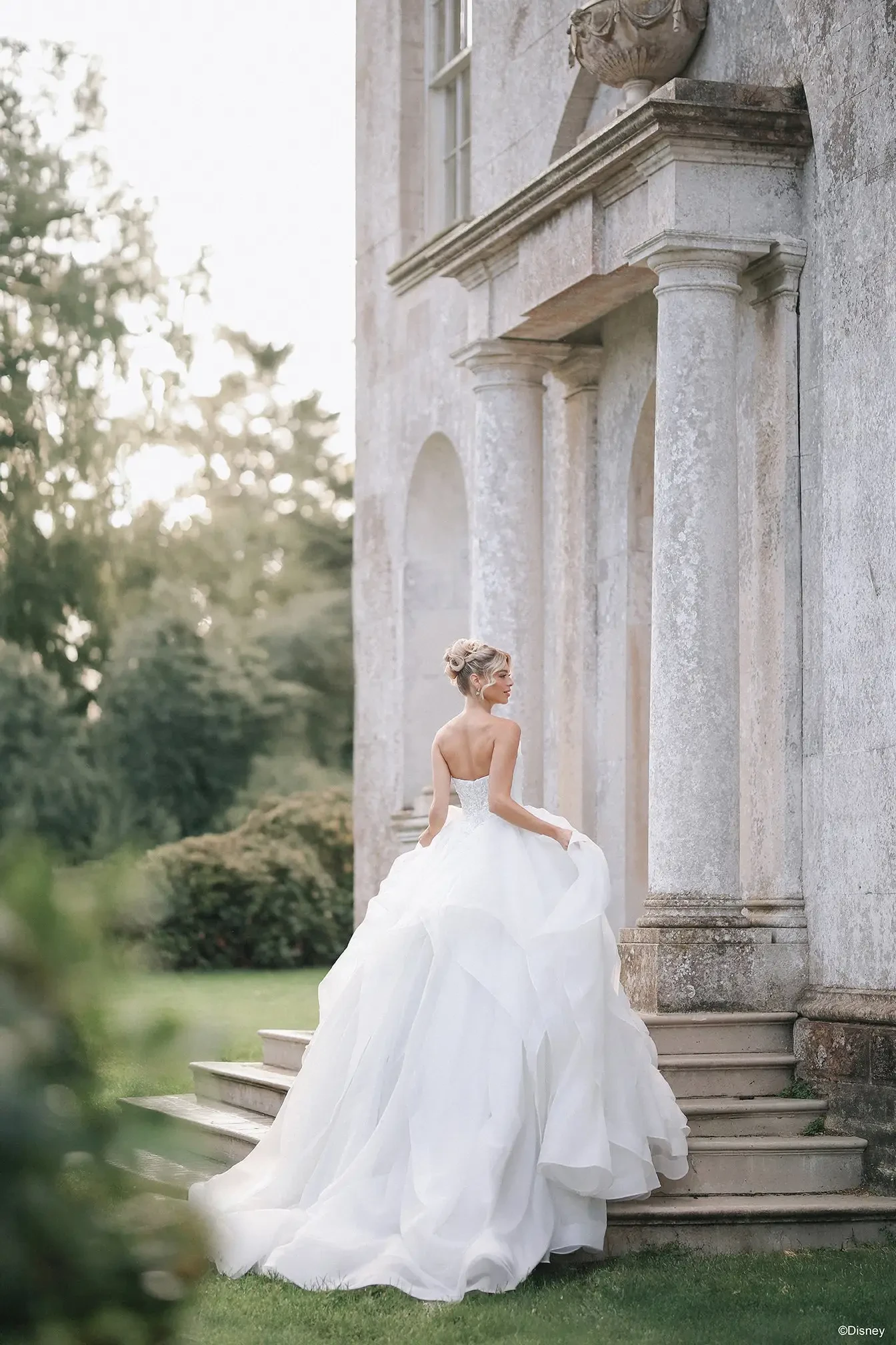 A woman in a white wedding dress stands on the steps of a grand building, looking back over her shoulder. The setting is lush with greenery and soft sunlight.