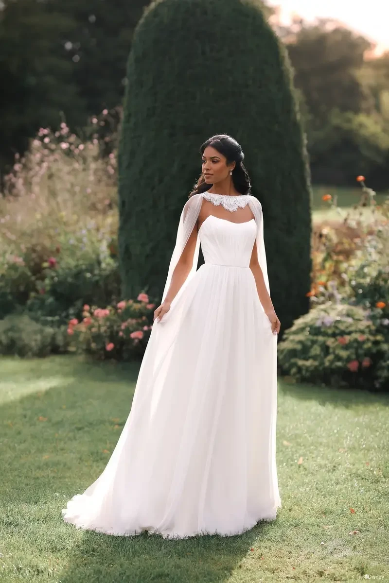 A woman in a white wedding dress with a cape stands in a lush garden, surrounded by flowers and greenery.