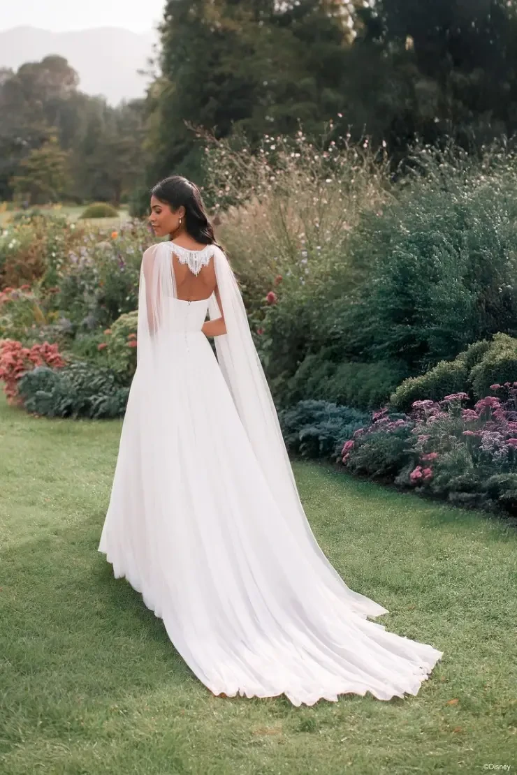A woman wearing a white gown with a flowing train and a cape stands in a garden, facing away from the camera. The background features greenery and colorful flowers.