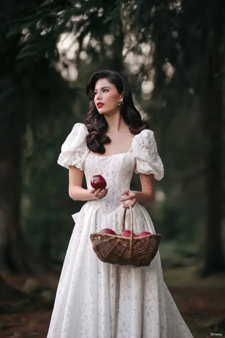 A woman in a white dress with puff sleeves stands in a forest, holding a basket full of apples and two red apples in her hands.
