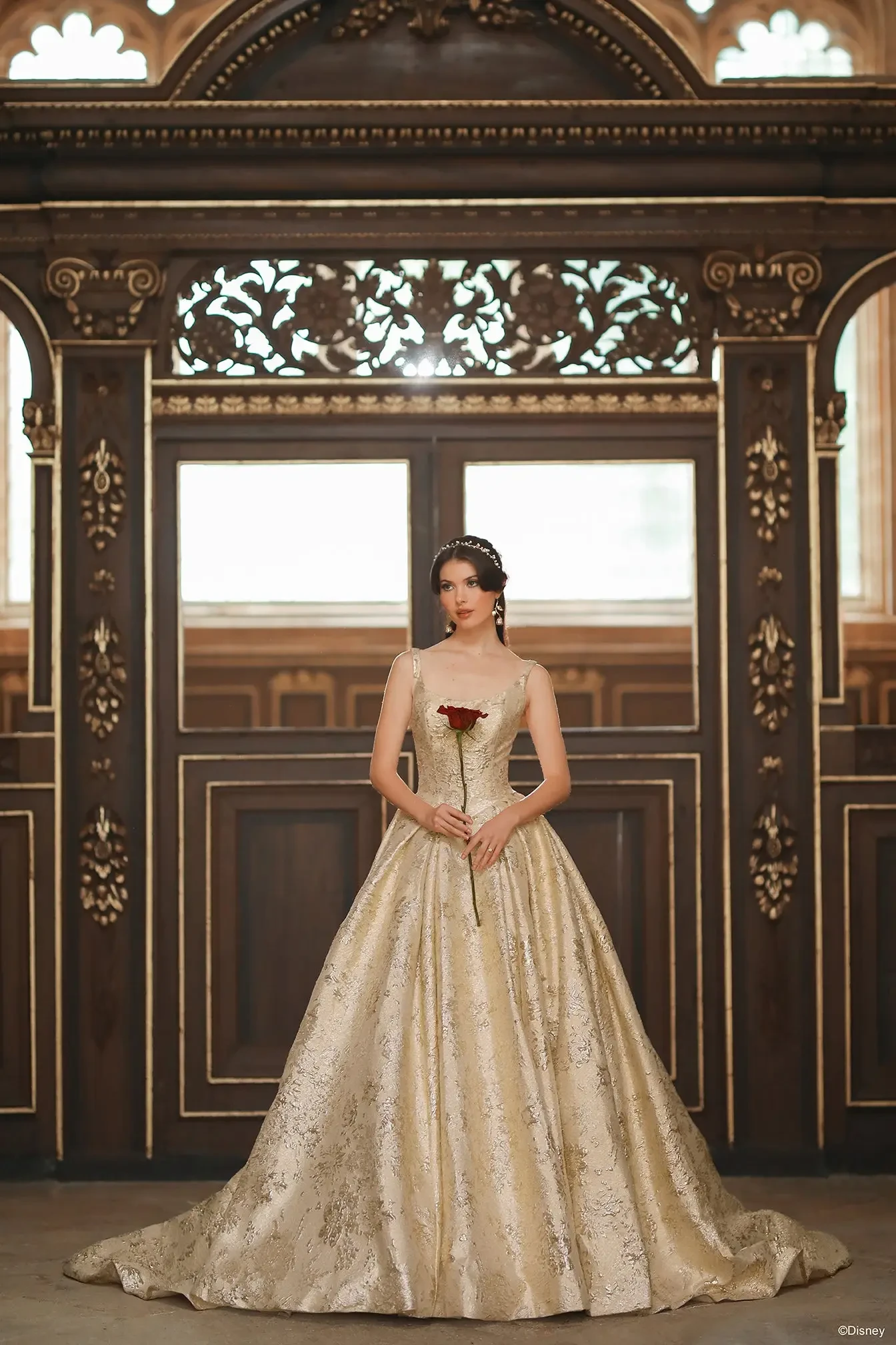 A woman in an elegant, gold-colored ball gown with intricate patterns stands in front of ornate wooden architecture, holding a single red rose.
