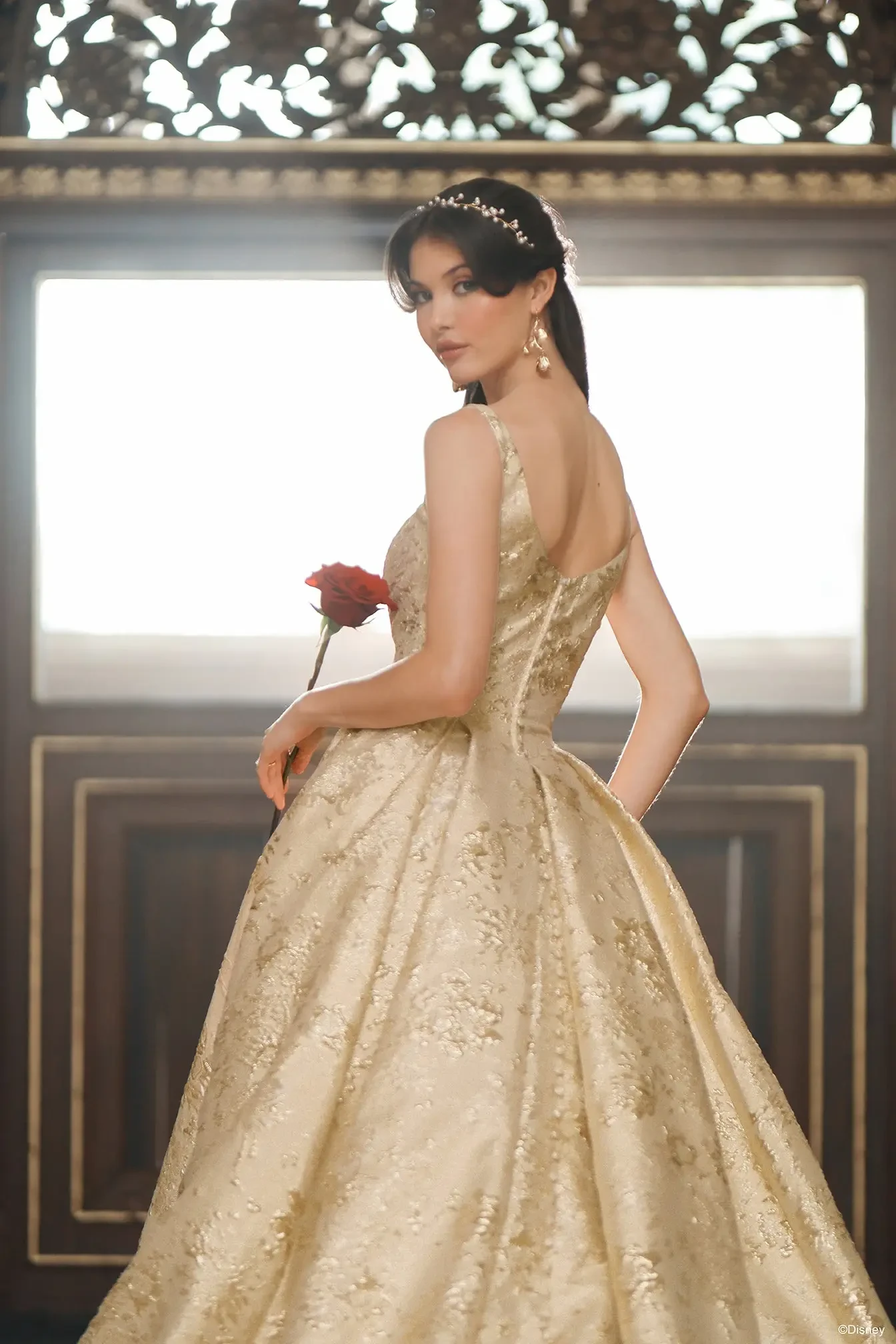 A woman in an elegant golden gown holds a red rose and poses elegantly with her back turned, showcasing intricate details of the dress. The background features ornate architecture and soft lighting.