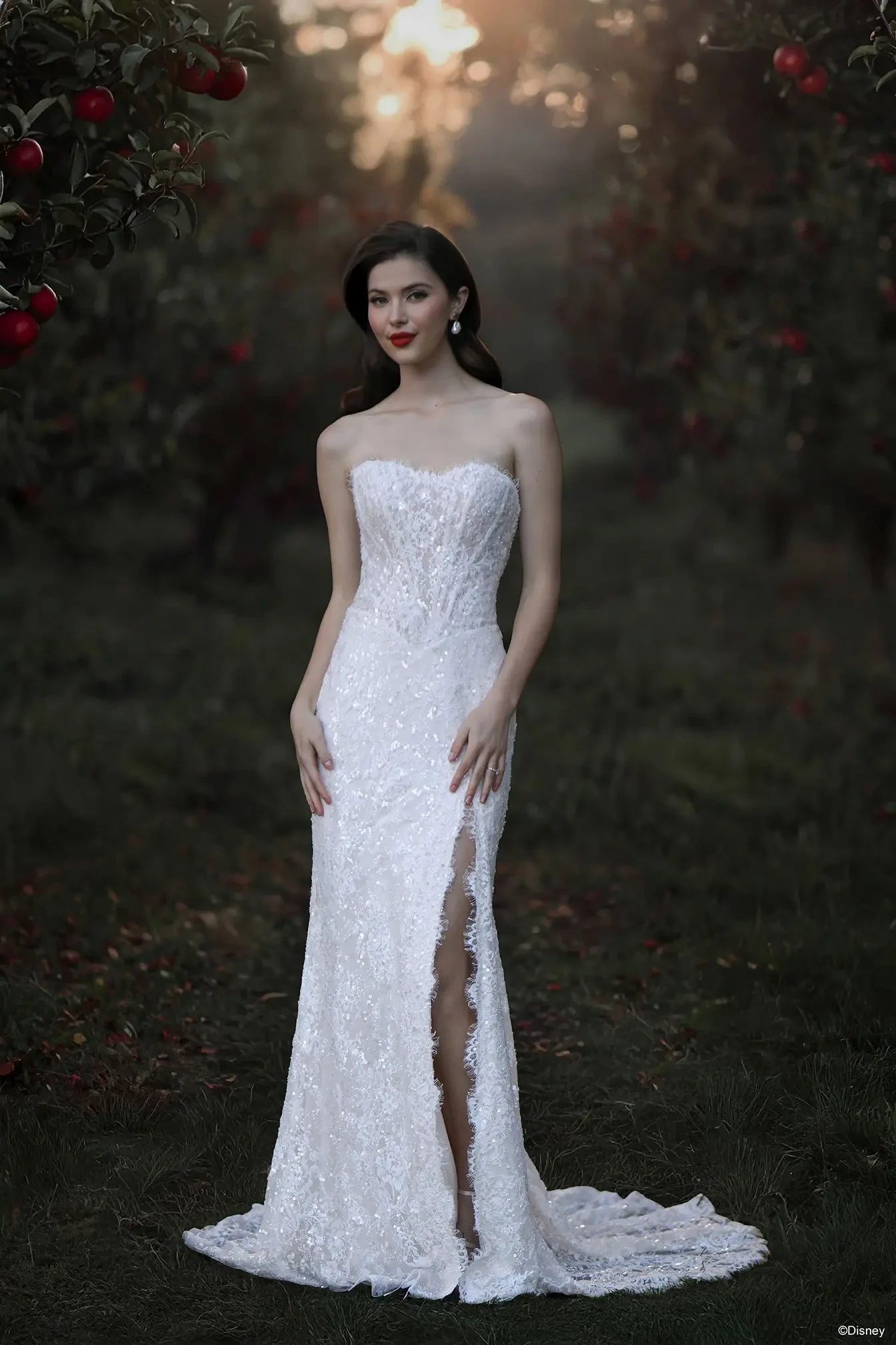 A woman in a strapless white gown with intricate detailing and a thigh-high slit, standing in an orchard with red apples and soft sunlight filtering through the trees.