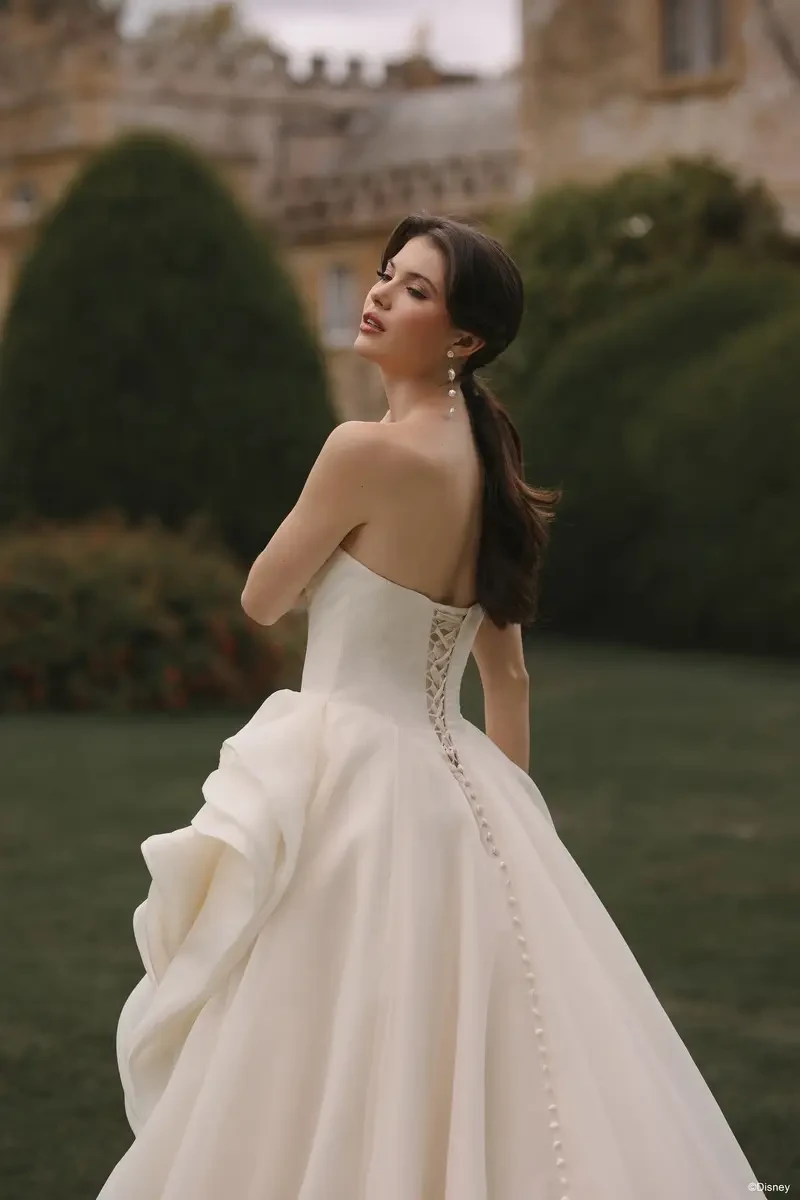 A woman in a strapless white wedding dress with a flowing skirt and buttons down the back, standing outdoors with her back to the camera and looking over her shoulder.