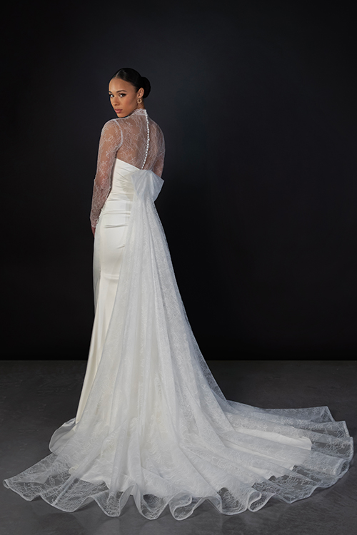 A woman in a sleek, long white gown with a lace back poses against a dark background. The dress features a flowing train, conveying elegance and sophistication.