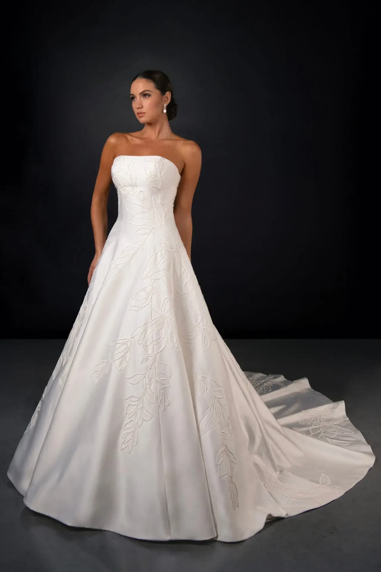 A woman in a strapless, white wedding gown with a flowing train, standing against a dark background.