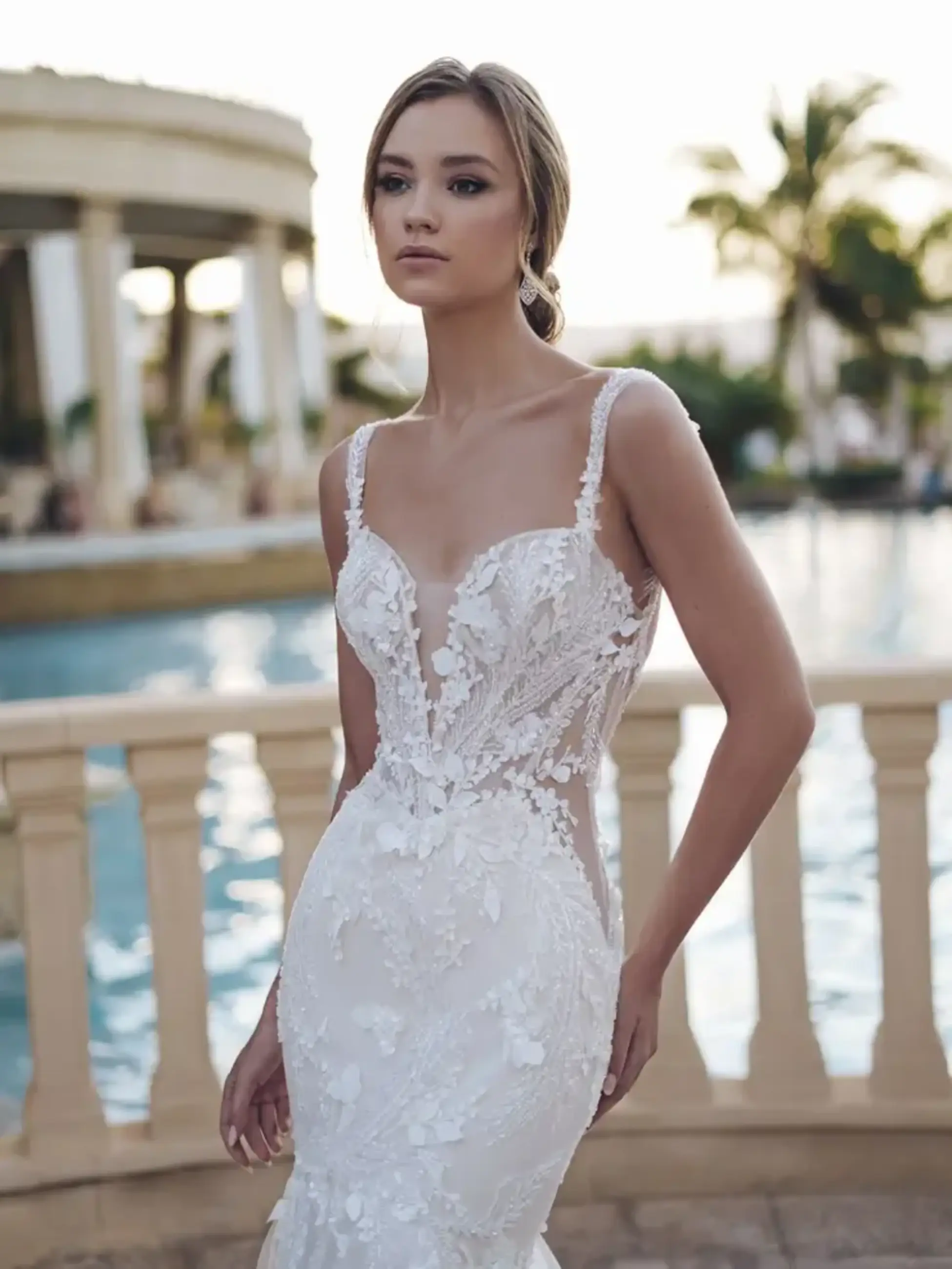A bride stands outdoors in an elegant lace wedding gown with a mermaid silhouette. Behind her, a pool and palm trees create a serene, tropical ambiance.
