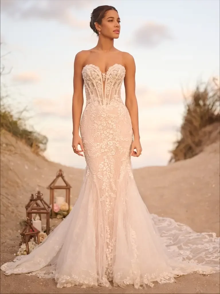 A woman in a fitted, strapless wedding dress with intricate lace details, standing on a sandy pathway surrounded by grass and lanterns.