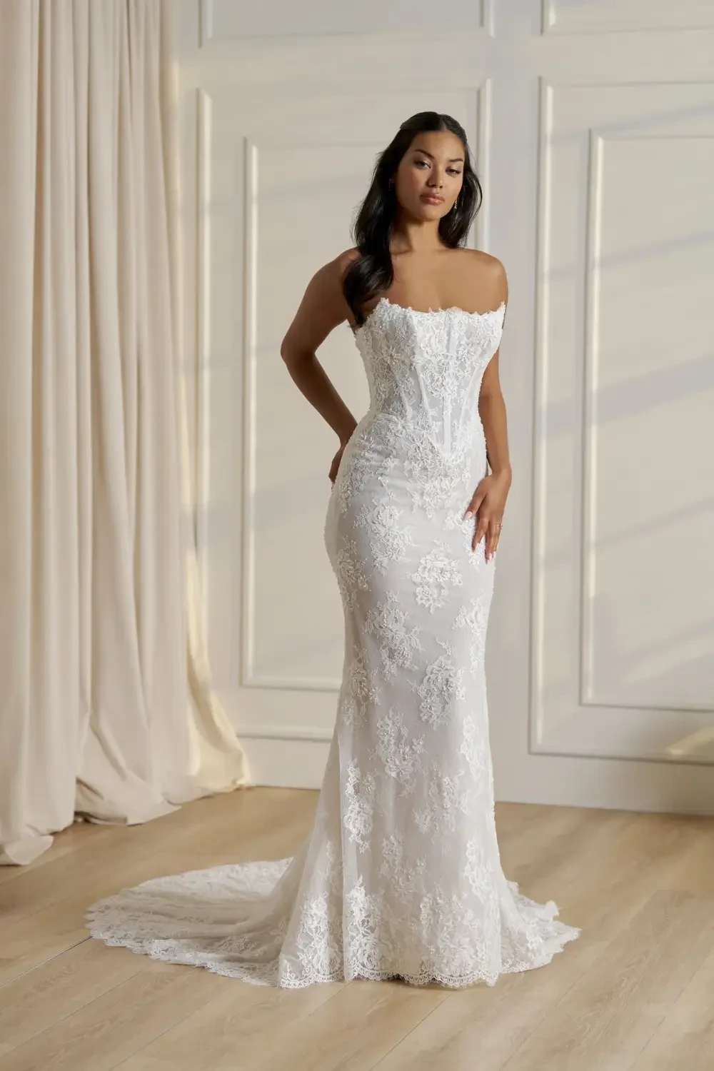 A woman wearing a fitted white lace wedding dress with off-the-shoulder sleeves, standing gracefully in an elegant indoor setting.