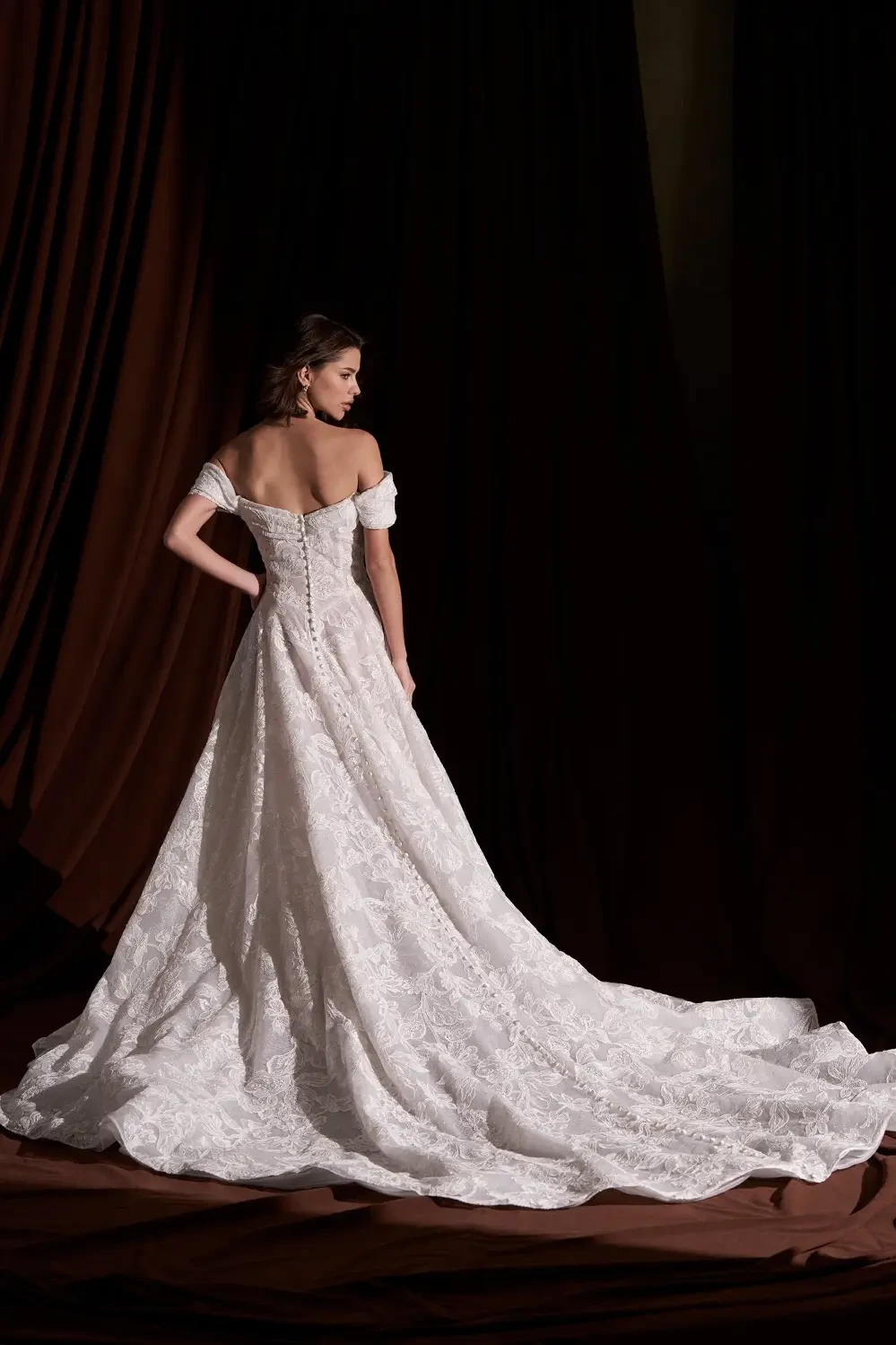 A model wearing a strapless, off-the-shoulder white lace gown, standing against a dark brown background.