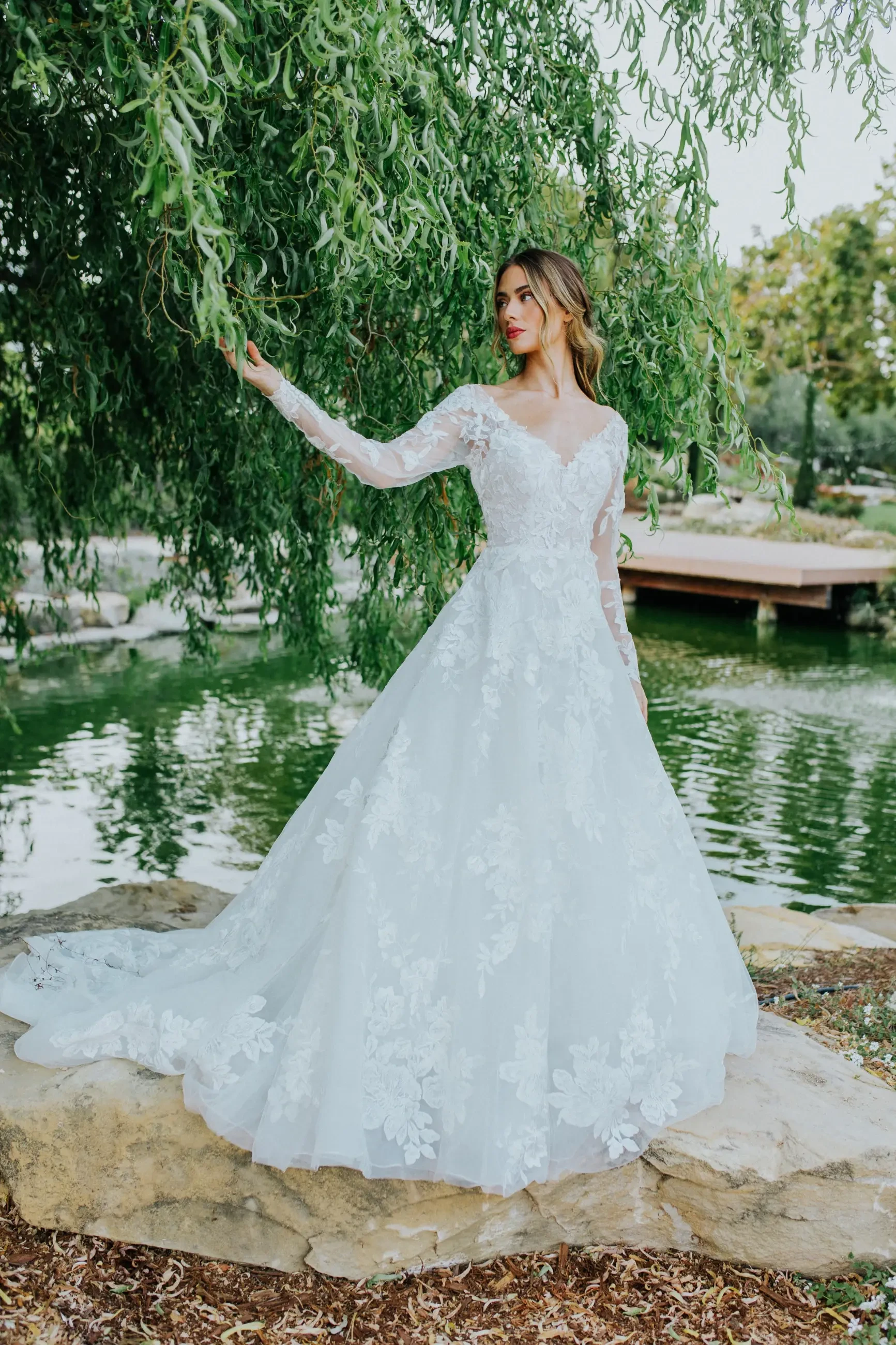 A bride in a lace wedding gown stands by a serene pond under a willow tree, exuding elegance and tranquility. Stone and greenery surround her.