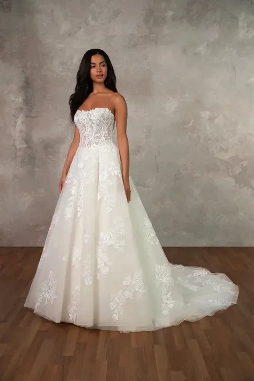 A woman in a strapless white wedding dress with intricate floral embroidery standing on a wooden floor against a textured gray background.