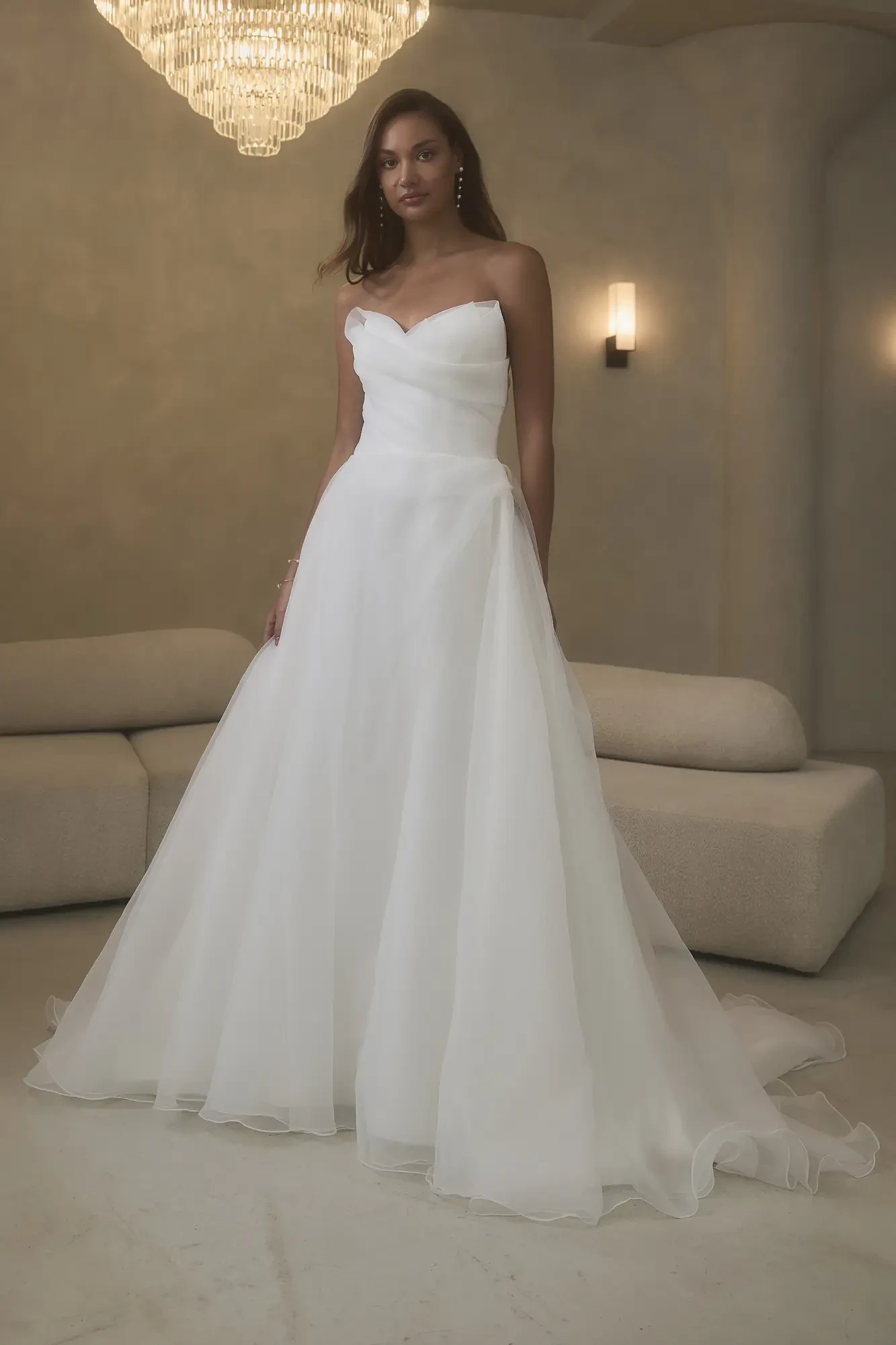 A woman wearing a strapless white wedding gown stands elegantly in a well-lit room.