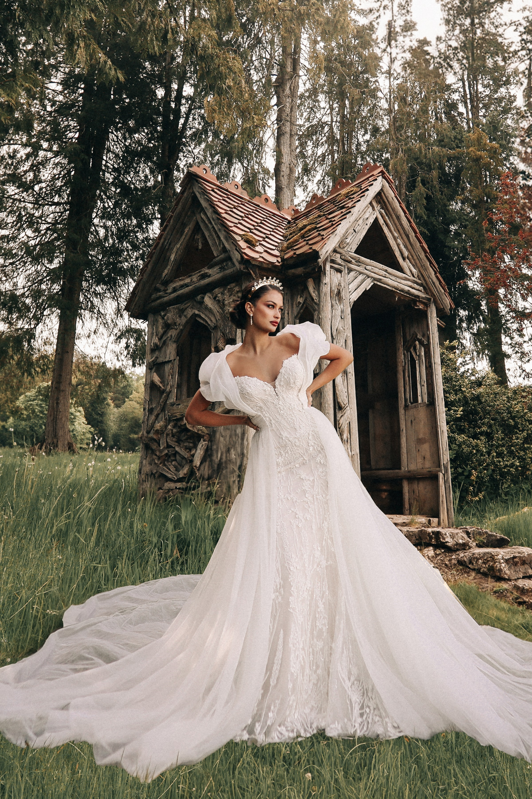 A woman in an elegant wedding dress stands confidently outside a rustic wooden cottage surrounded by greenery.