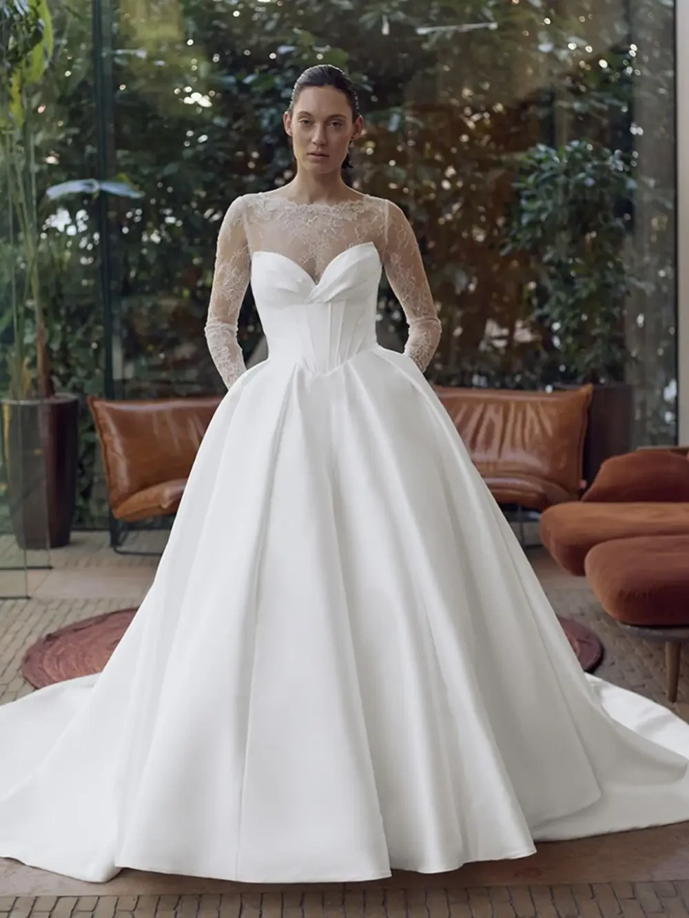 A woman in a sophisticated white gown with a fitted bodice and a full skirt, standing in a stylish indoor setting with green plants and modern furniture.
