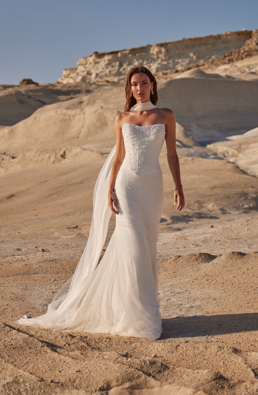 Bride in a strapless, flowing white gown stands on sandy terrain, under clear blue sky, with a serene and confident expression.