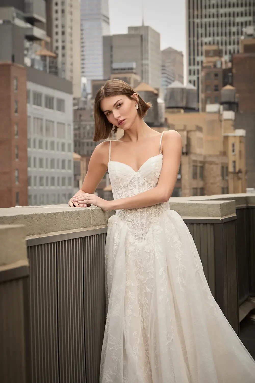 A model poses in a wedding dress on a rooftop with a city skyline in the background.