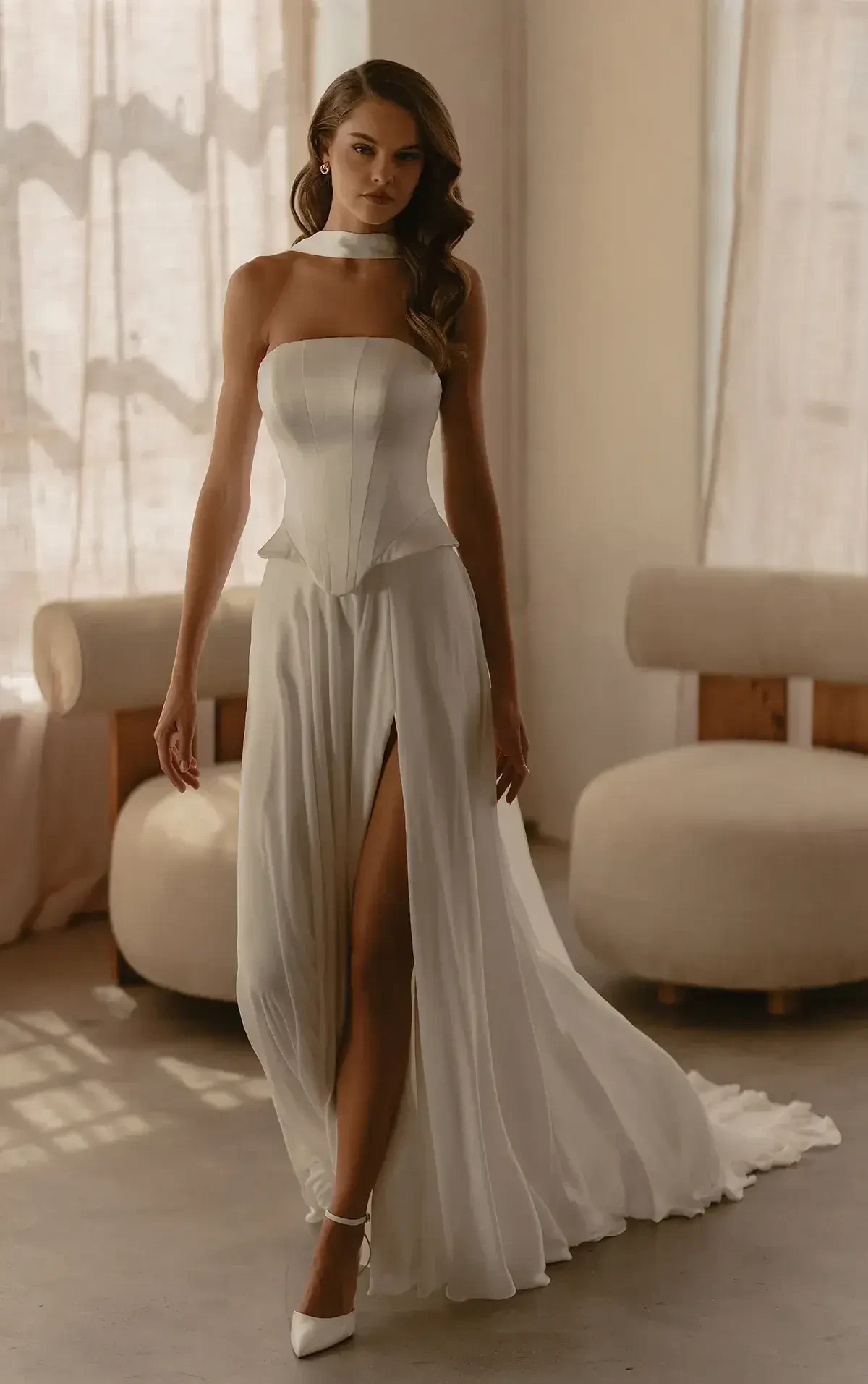 A woman in an elegant white strapless gown with a flowing skirt and high slit poses by a window. The room's soft lighting creates a serene mood.