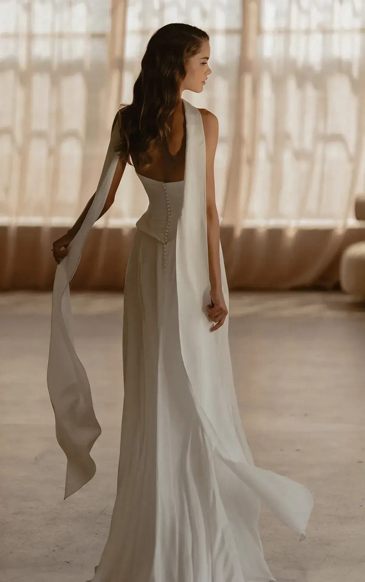 A woman in an elegant white strapless gown with a flowing skirt and high slit poses by a window. The room's soft lighting creates a serene mood.