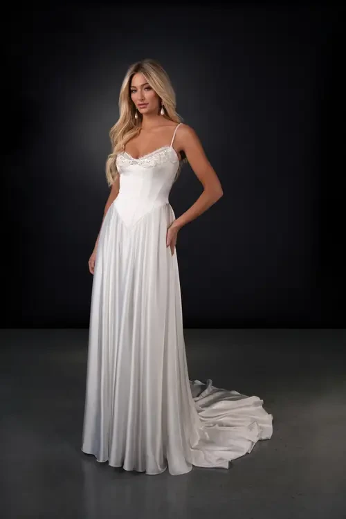 A model wearing a white gown with a slight train, standing against a dark background.