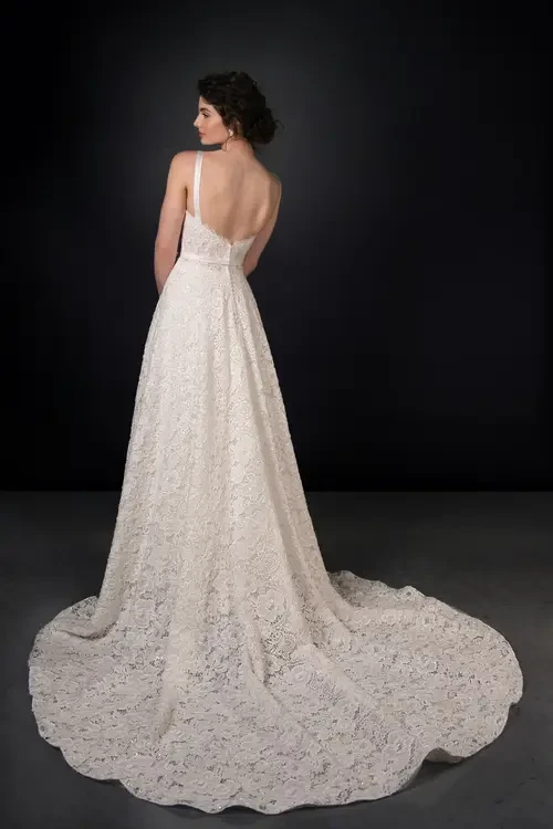 A model wearing a backless, white lace wedding dress with a long train, standing against a dark background.