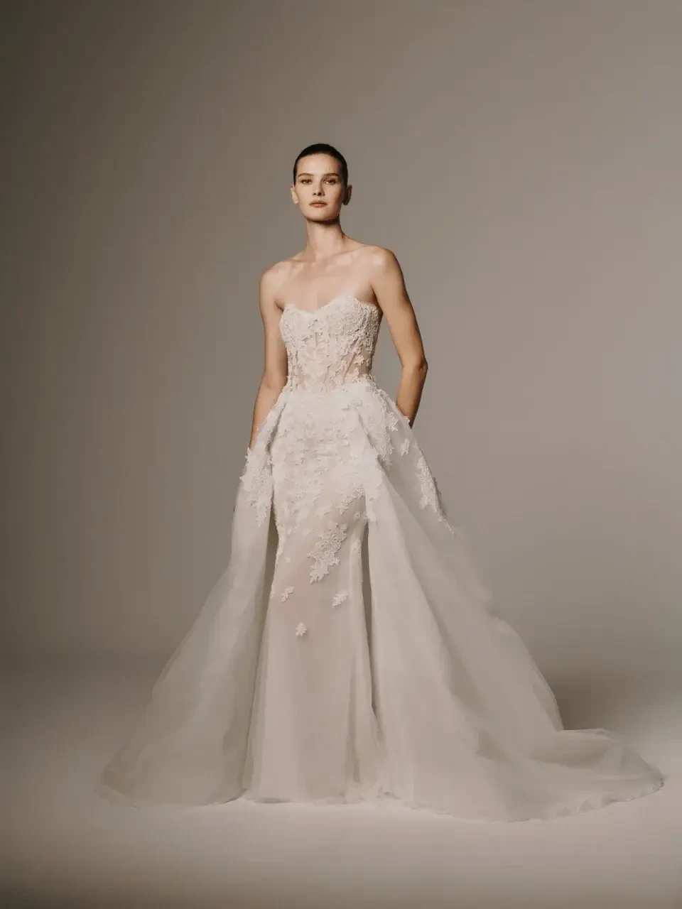 A model wearing a strapless wedding gown with intricate detailing and a flowing train. The gown features floral embellishments and is set against a neutral background.