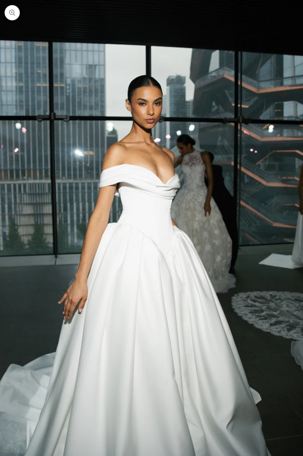 A model poses in a stunning white off-the-shoulder gown with a full skirt, showcasing a sophisticated and elegant style.