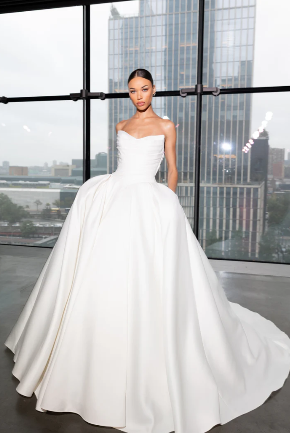 A model in a strapless white gown poses in a modern building with large windows, showcasing a city skyline in the background.