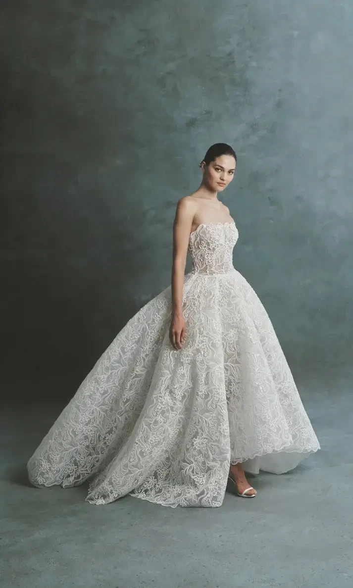 A model wearing a strapless, intricately designed white gown with a voluminous skirt, standing against a textured blue-green background.