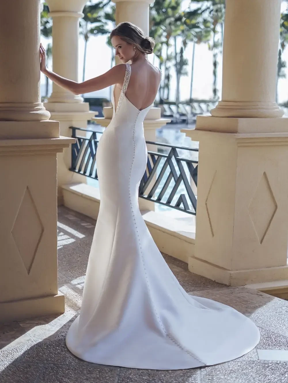 A woman in a sleek, elegant wedding dress poses with her back to the camera, showcasing the dress's fitted silhouette and button details, with a scenic background of palm trees and water.