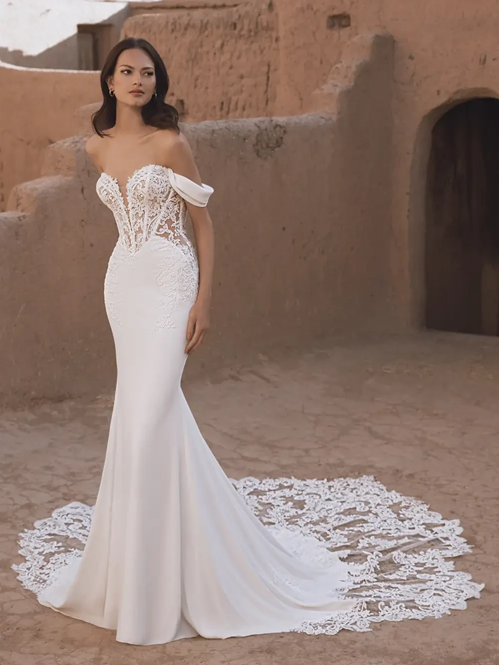 A woman in an elegant, fitted white wedding dress with lace details, standing outside against a textured earth-toned backdrop.